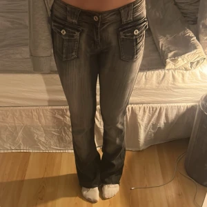 Grå bootcut jeans med stora fickor - Säljer ett par grå jeans med bootcut-snitt och stora fickor med knappstängning fram och bak. Byxorna har låg midja och coola slitningar som ger en snygg streetkänsla. Perfekt för dig som gillar detaljer och vill sticka ut.
