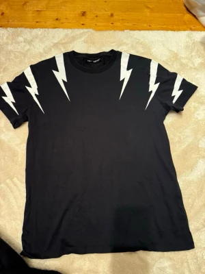 Svart Neil Barrett t-shirt - Säljer en svart t-shirt från Neil Barrett med vita blixtmotiv över axlarna och ärmarna. Nypris 2499
