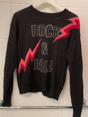 Svart stickad tröja Zadig & Voltaire - Cool svart stickad tröja från Zadig & Voltaire med texten 'Rock & Roll' framtill och två röda blixtar som ger en edgy vibe. Rund halsringning och långa ärmar. 