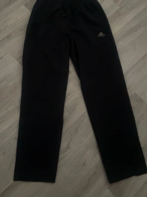 Svarta träningsbyxor från Adidas - Svarta träningsbyxor från Adidas med diskret logga på vänster lår. Byxorna har rak passform och elastisk midja med snörning för extra komfort. Materialet är mjukt och passar perfekt för sport eller chill. Snygg och enkel design som funkar till många stilar. Jackan-200 byxan-250