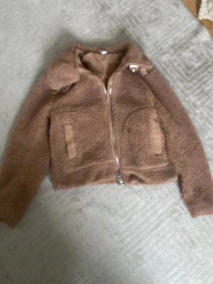 Brun teddyjacka med dragkedja - Mysig brun teddyjacka med croppad passform och silverfärgad dragkedja framtill. Jackan har två fickor och en klassisk krage. Perfekt för dig som gillar en chill och trendig look under höst och vinter.