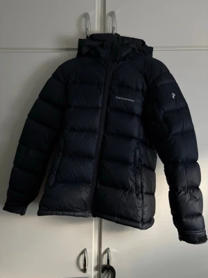 Svart Peak Performance dunjacka - Säljer en svart dunjacka från Peak Performance med huva och dragkedja framtill. Jackan har ett puffigt quiltat utseende och logga på bröstet och ärmen. Perfekt för kalla dagar och har en clean, sportig vibe. Materialet är syntet och dun, och jackan är långärmad.