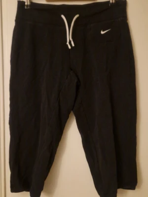 Svarta Nike Dri-Fit mjukisbyxor M - Svarta knälånga mjukisbyxor från Nike med Dri-Fit-teknologi och vit snörning i midjan. Snyggt broderad Nike-logga på fickan och ett extra resårband på insidan av linningen. Sköna och funktionella för träning eller chill. Passar dig som gillar sportig stil.