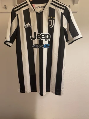 Juventus adidas fotbollströja - Juventus hemmatröja från adidas med klassiska svarta och vita ränder, broderat klubbmärke med tre guldstjärnor och Jeep-logga på bröstet. Tröjan har rund hals och är tillverkad i Primegreen-polyester, perfekt för fotboll och träning.