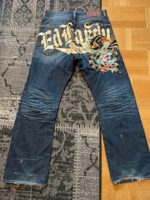 Ed Hardy blå jeans med tryck - Fetaste ed Hardy jeansen. Flared och unik design på baksidan. Storlek 34/32.               Längd: (110cm).  Midja (45cm).  Över låren (31cm). Benöppningen (25,5cm)