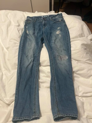 Blå jeans från Here's To The Boys - Säljer ett par blå jeans från Here's To The Boys med slitna detaljer och patch på ena benet. Klassisk femficksmodell med raka ben och normal passform. Jeansen har en snygg tvätt och är gjorda i bomullsmaterial.