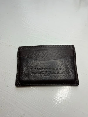Barbour mörkbrun korthållare i skinn - Korthållare från Barbour i mörkbrunt skinn. Flera kortfack på båda sidor och ett öppet fack i mitten med klassiskt rutigt innerfoder.