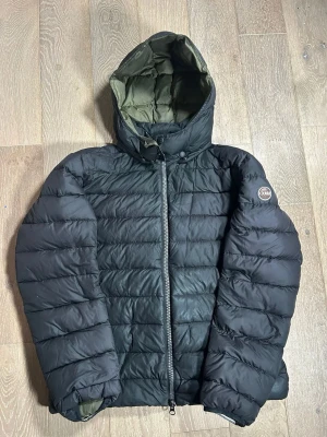 Svart pufferjacka från Colmar - Snygg svart pufferjacka från Colmar med huva och grönt foder. Jackan har en rak passform, quiltad design och dragkedja framtill. Perfekt för kalla dagar och har en cool badge på ärmen. Passar dig som gillar stilren och funktionell vinterjacka.