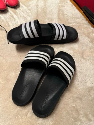Svarta Adidas sandaler med vita ränder - Säljer ett par klassiska svarta Adidas sandaler med tre vita ränder över foten. Modellen har öppen tå och platt sula, perfekt för chill dagar. Tillverkade i syntetmaterial och har en enkel, sportig look som funkar till allt.