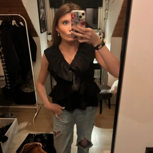 Svart omlott blus med volang - Säljer en svart omlott korsad blus med volangdetaljer och djup v-ringning. Blusen är ärmlös och har en snygg, lite utsväng passform som ger en cool siluett. Materialet känns mjukt och har en lätt glans. Perfekt att styla med jeans eller kjol för en trendig look.