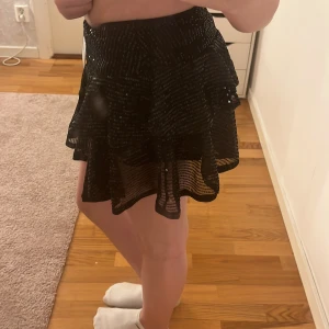 Svart paljettkjol med volang - Säljer en svart kortkjol med glittriga paljetter och volangdetaljer. Kjolen har inbyggda shorts. Aldrig använd. Strl S