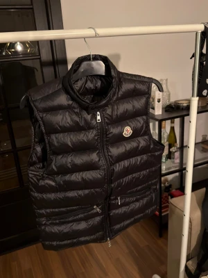 Svart dunväst från Moncler Gui - Säljer en svart dunväst från Moncler med quiltad design och hög krage. Västen har två praktiska fickor med dragkedja och ett broderat Moncler-märke på bröstet. Materialet är glansigt och känns lätt men värmande, perfekt för lager-på-lager. Dragkedja framtill för enkel stängning. I princip helt ny! Bästa batchen!