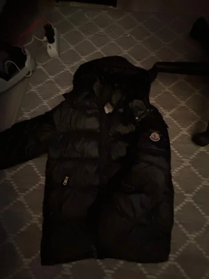 Svart Moncler Maya - Säljer en svart pufferjacka från Moncler med huva och coolt Moncler-märke på ärmen. Jackan har dragkedjor på fickorna och quiltad design som ger en schysst look. Insidan har en unik serietidningsetikett. Perfekt för dig som vill ha en varm och snygg jacka.