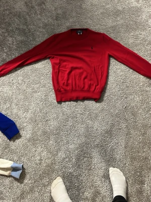 Röd tröja från Polo Ralph Lauren - Snygg röd tröja från Polo Ralph Lauren med klassisk rund hals och diskret broderad logga på bröstet. Tröjan har långa ärmar och ribbade muddar vid ärmslut och nederkant. Perfekt för dig som gillar stilrena och tidlösa plagg.
