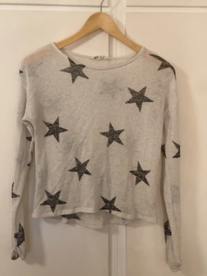 Vit stickad tröja med svarta stjärnor H&M - Vit stickad tröja från H&M med svarta stjärnor över hela plagget. Tröjan har rund halsringning och långa ärmar. Materialet är mjukt och lätt, perfekt för att ge en chill vibe till din outfit.