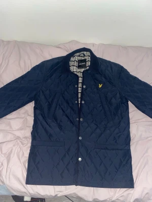 Mörkblå quiltad jacka från Lyle & Scott - Quiltad mörkblå jacka från Lyle & Scott med tryckknappar och klassisk krage. Insidan har ett rutigt foder i vitt och svart. Jackan har två stora fickor framtill och den ikoniska gula logon på bröstet. Perfekt för dig som vill ha en stilren och bekväm look.