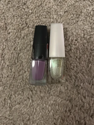 Isadora nagellack duo lila & glitter - Två nagellack från Isadora: ett i lila nyans med svart kork och ett skimrande glitterlack med vit kork. Båda har bred pensel för enkel applicering och kommer i rektangulära glasflaskor. Perfekt för att mixa och matcha färger och effekter på naglarna.