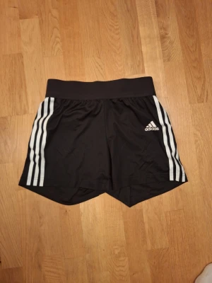Svarta Adidas träningsshorts XS - Säljer ett par svarta Adidas shorts i storlek XS med vita ränder längs sidorna och logga på framsidan. De har bred resår i midjan och snörning på insidan. Materialet är lätt och andas, perfekt för sport och träning.