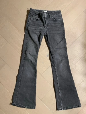 Bootcut jeans från Gina Tricot, grå - Snygga grå bootcut jeans från Gina Tricot i storlek 146. Klassisk femficksmodell med dragkedja och knapp framtill. Jeansen har en mörkgrå tvätt och är tillverkade i mjukt denim-material som sitter skönt. Perfekt för dig som gillar retrovibbar och vill ha en cool siluett.