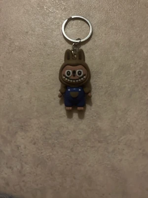 Ingen väst på nyckelring - Bilderna visar en nyckelring med en figur i blå overall och brunt kaninhuvud, men ingen väst syns på figuren. Ingen relevant information om väst kan tas ut.