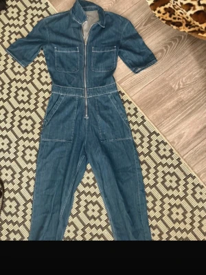 Blå jeans jumpsuit från ONLY, strl 34 - Säljer en snygg blå jeans jumpsuit från ONLY i storlek 34. Modellen har korta ärmar, två stora bröstfickor och dragkedja framtill. Materialet är klassisk denim med vita sömmar och jumpsuiten har en rak passform med markerad midja. Straight fit, aldrig använd
