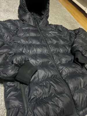 Svart Peak Performance dunjacka - Säljer en svart dunjacka från Peak Performance med huva och ribbade muddar. Jackan har en snygg quiltad design, dragkedja framtill och logga broderad på bröstet och ärmen. Perfekt för kalla dagar och har en modern, loose passform.