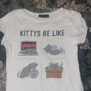 Vit t-shirt med kattmotiv från FB Sister - Säljer en vit t-shirt från FB Sister i storlek XS med trycket 'KITTYS BE LIKE' och fyra söta kattillustrationer. T-shirten har rund hals och korta ärmar. Materialet är mjuk bomull och motivet är färgglatt med inslag av rött, blått och grått.