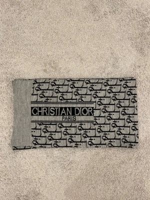 Christian Dior Paris halsduk - Snygg stickad halsduk från Christian Dior Paris med grå och svart färg. Halsduken har ett grafiskt mönster med Dior-loggor över hela och en bred logotyp framtill. Materialet är mjukt och stickat, perfekt för att ge din outfit en lyxig touch.