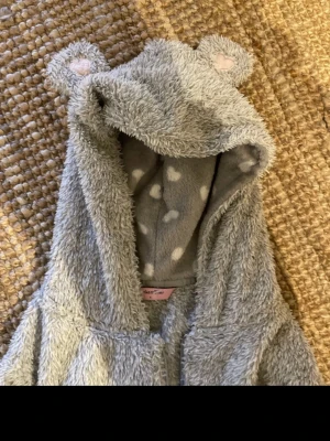 Grå teddyjacka med öron från SmåkerSister - Supermysig grå teddyjacka med huva och söta öron med hjärtan. Insidan av huvan har ett gulligt hjärtmönster i vitt. Jackan har dragkedja framtill och ribbade muddar vid ärmslut. Perfekt för chill dagar eller när du vill vara extra cozy.