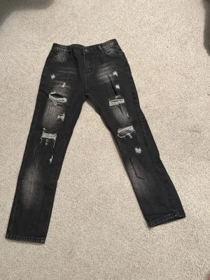Svarta ripped jeans, storlek 14Y - Säljer ett par svarta jeans med slitna detaljer och stora hål på benen. Jeansen har en skinny passform och är tillverkade i klassiskt denimtyg. Perfekta för dig som gillar en edgy och trendig stil.