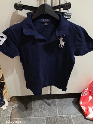Mörkblå pikétröja från Polo Ralph Lauren - Säljer en mörkblå pikétröja från Polo Ralph Lauren med klassisk krage och korta ärmar. Tröjan har en stor vit broderad logga på bröstet och en vit siffra på ena ärmen. Materialet är mjuk bomull och modellen är normal passform.