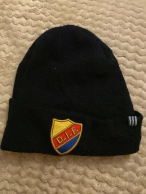 För 13-15 år typ Svart DIF mössa med broderad logga - Svart mössa med broderad DIF-logga framtill i gult, rött och blått. Klassisk design med uppvikt kant och diskreta ränder på sidan. Perfekt för dig som vill visa ditt lagstöd.