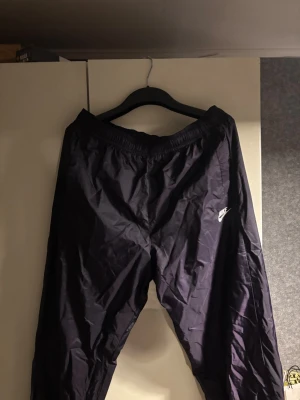 Svarta Nike täckbyxor XL - Nike täckbyxor i svart med elastisk midja och meshfoder. Byxorna är liten i storleken den passar mer L än Xl. Diskret Nike-logga på benet och enkel design utan extra detaljer
