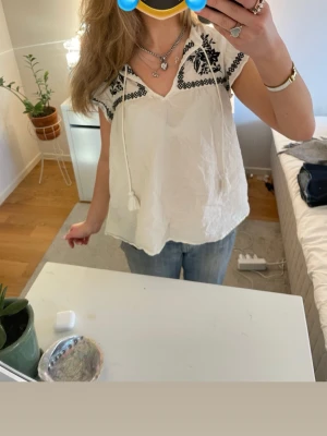 Vit blus med svart broderi och knyt - Säljer en vit blus i linne med svart broderat blommönster vid axlarna och halsen. Blusen har korta ärmar, v-ringning och dekorativ knytning med tofsar framtill. Materialet är lätt och luftigt, perfekt för varmare dagar.