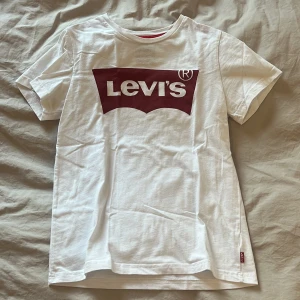 Vit Levi's t-shirt med röd logga - Säljer en klassisk vit t-shirt från Levi's i storlek 12. Tillverkad i 100% bomull, är tjock i bra kvalitet och icke-genomskinlig. Storlek 12 motsvarande 152cm.