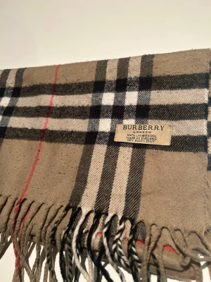 Rutig beige halsduk från Burberry - Säljer en klassisk rutig halsduk från Burberry i beige, svart, vitt och rött. Halsduken har det ikoniska Burberry-mönstret och är tillverkad i mjuk ull. Perfekt för dig som vill ha en tidlös och snygg look med lite lyxig känsla.
