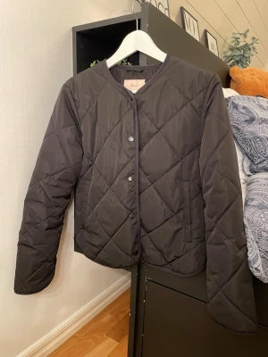 Svart quiltad jacka från Nelly - Quiltad svart jacka från Nelly i storlek S. Jackan har rund hals, tryckknappar framtill och ett stilrent rutmönster. Perfekt för dig som gillar en clean look med lite edge. Materialet är syntet och jackan har långa ärmar.