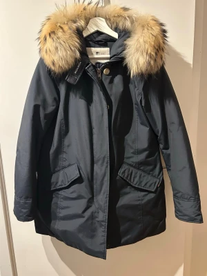 Woolrich Arctic Parka, storlek M - Woolrich Arctic Parka i mörkblå färg med fluffig päls runt huvan. Jackan har både dragkedja och knappar framtill. Mycket fint skick då den är väldigt sparsamt använd. Minimalt slitage. Storlek M.