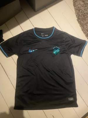 Svart Nike fotbollströja Brasilien - Säljer en svart Nike fotbollströja med Brasilien-emblem och fem stjärnor på bröstet. Tröjan har blå detaljer vid krage och ärmslut samt ett blått Nike Swoosh på framsidan. Materialet är lätt polyester som andas och passar perfekt till träning.