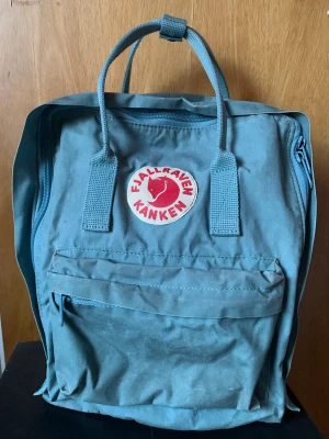 Blå Fjällräven Kånken ryggsäck - Säljer en klassisk gråblå Fjällräven Kånken ryggsäck med det ikoniska röda och vita logotypmärket framtill. Väskan har ett stort huvudfack, en mindre ficka med dragkedja på framsidan och justerbara axelremmar. Lite sliten och svagt missfärgad men hel och ren och har mycket kvar att ge. 