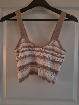 Beige och vit stickad linnetopp - Säljer en superfin stickad linnetopp i beige och vitt med breda axelband. Toppen har ett snyggt hålmönster och är croppad med vågig kant nedtill. Perfekt för varma dagar och ger en chill vibe till din outfit.