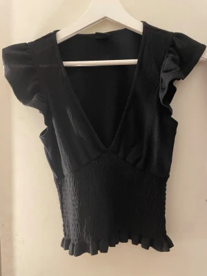 Svart blus med volangärm och v-ringning - Säljer en svart blus med djup v-ringning, volangärmar och smockad midja. Toppen har en croppad passform och volangdetaljer längst ner. Materialet är mjukt och lätt, perfekt för att lyfta din outfit.