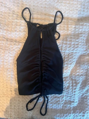 Svart ribbad linnetopp med snörning (Centerfold top från Chiquelle) - Snygg svart ribbad linnetopp (Centerfold top) med smala axelband och snörning både fram och bak. Toppen är croppad och har en åtsittande passform med rynkade detaljer framtill. Perfekt för att skapa en trendig look. Bortklippt lapp.