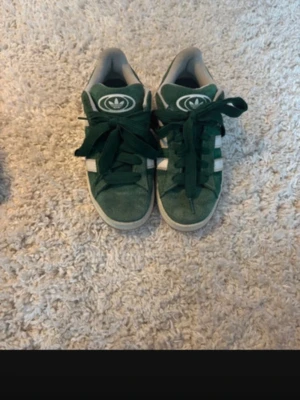 Adidas Campus gröna sneakers - Säljer ett par gröna Adidas Campus sneakers med klassiska vita ränder på sidorna och Adidas-logga på plösen och hälen. Skorna har breda gröna snören, rund tå och platt sula. Ovandelen är i mocka och insidan är ljusgrå. Perfekt för dig som gillar retrostil.🍃 OBS bilderna är från min Vinted, därav något suddiga! 
