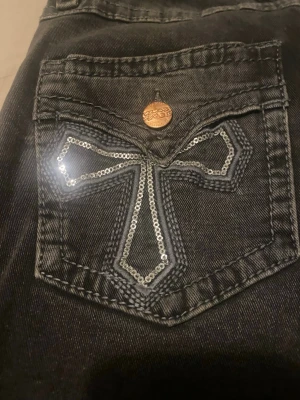 Svarta low-waisted jeans  - Säljer ett par svarta jeans med snygga paljettbroderade kors på bakfickorna och kopparfärgade knappar. Jeansen har en rak passform och är tillverkade i klassiskt jeansmaterial. Perfekt för dig som vill sticka ut med coola detaljer.