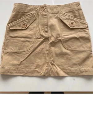 Beige kortkjol med fickor och knappar - Säljer en beige kortkjol i bomull med två stora fickor framtill, dekorerade med knappar. Kjolen har en rak passform och stängs med knapp och dragkedja framtill. Perfekt till sommaren och enkel att matcha med olika toppar. Aldrig använd 