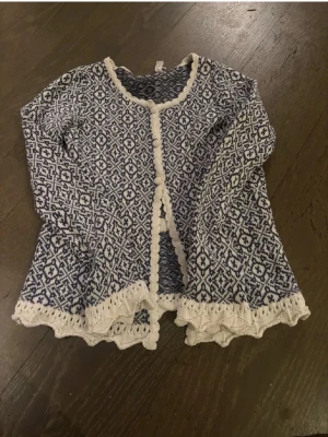 Blåvit mönstrad kofta med volangkant - Supersöt kofta från Rosebud i blått och vitt med detaljerat stickat mönster. Koftan har volangkant, dekorativa knappar framtill och lång ärm. Fina virkade kanter i vitt ger en extra touch. Perfekt för dig som gillar unika plagg med vintagekänsla.