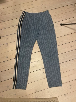 Blå mönstrade mjukisbyxor Lacoste M - Säljer ett par blå mjukisbyxor från Lacoste i storlek M. Byxorna har ett grafiskt mönster, vita och mörkblå sidoremsor samt den klassiska Lacoste-loggan broderad vid fickan. Resår i midjan och regular fit för skön passform.