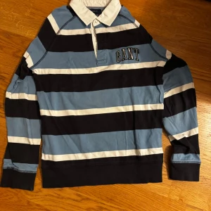 Randig långärmad skjorta från GANT - Snygg långärmad skjorta från GANT med breda blå, svarta och vita ränder. Klassisk krage och knappar framtill, samt broderad GANT-logga på bröstet. Perfekt för dig som gillar sportig och avslappnad stil.