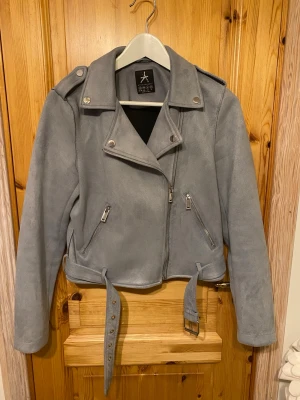 Grå bikerjacka mocka imitation  - Snygg grå bikerjacka i mocka imitation. Jackan har klassisk krage, silverfärgade dragkedjor, axelklaffar och bälte i midjan. Croppad modell med långa ärmar och två fickor med dragkedja framtill. Perfekt för dig som gillar stilrena detaljer.